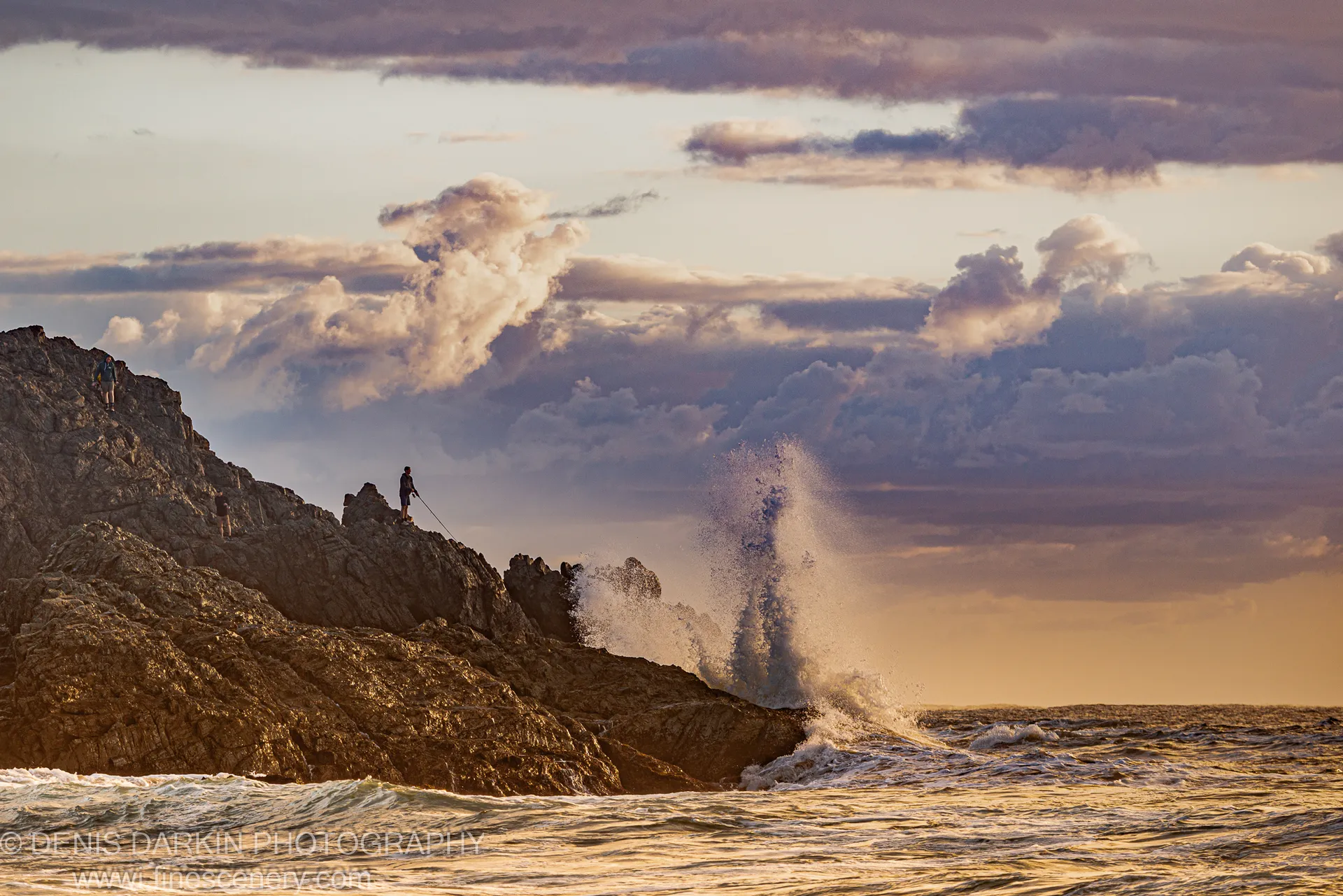 A column of white spray erupts from a wave striking the cliff base at Coffs Harbor, with violet and peach clouds above.