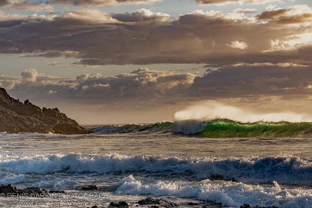 A large wave with an emerald crest explodes into white spray against dark rocks and a clouded sky at Coffs Harbor.