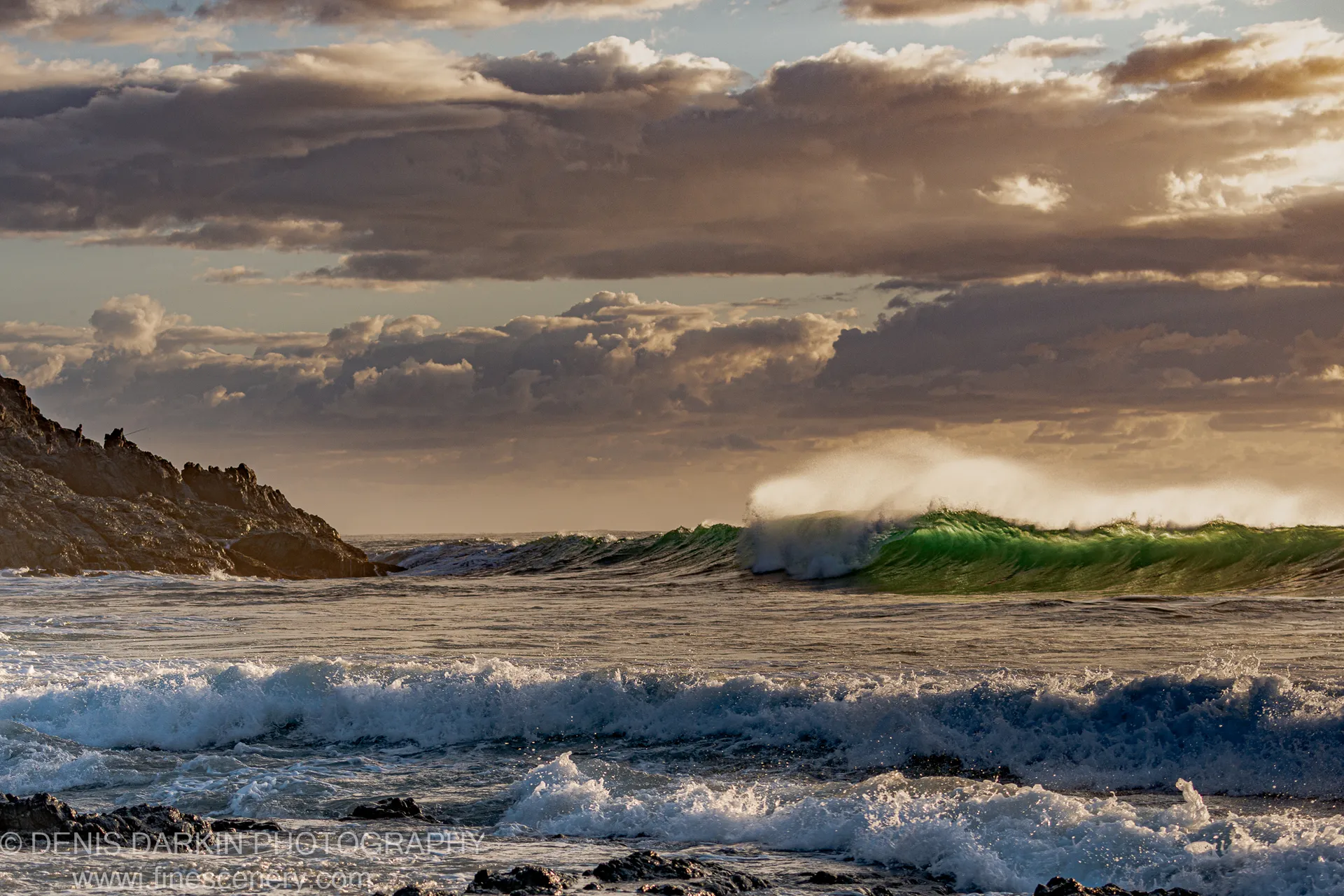A large wave with an emerald crest explodes into white spray against dark rocks and a clouded sky at Coffs Harbor.
