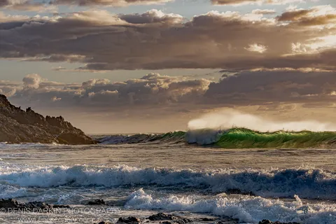 A large wave with an emerald crest explodes into white spray against dark rocks and a clouded sky at Coffs Harbor.