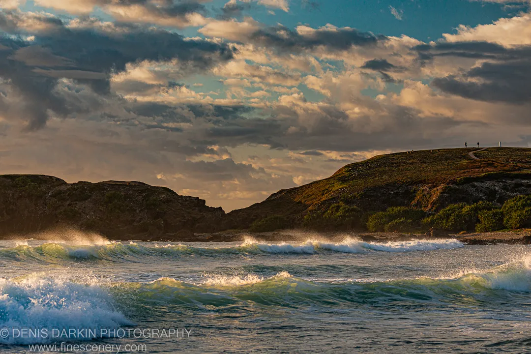 Small figures walk along a grassy headland at Coffs Harbor as waves roll across turquoise water toward a rocky foreshore.