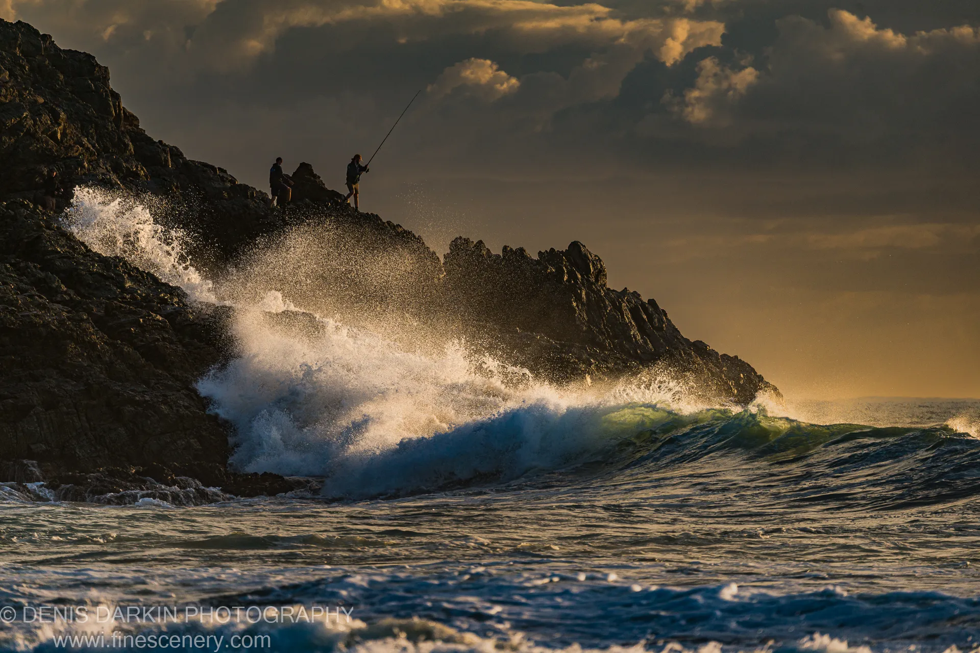Two rock fishers stand silhouetted on a jagged cliff at Coffs Harbor as a wave crashes below them in backlit spray.