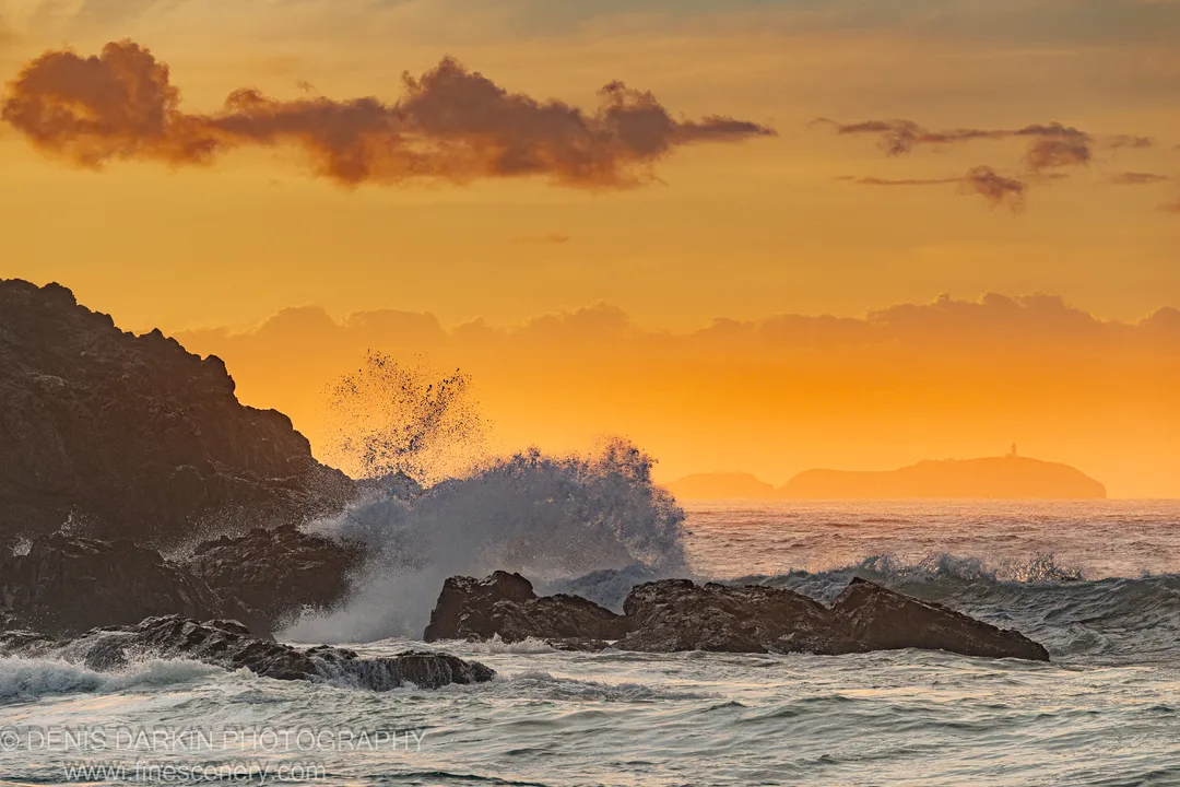 A wave explodes against a dark cliff face at Coffs Harbor, sending white spray into the air beneath an orange sunrise sky.