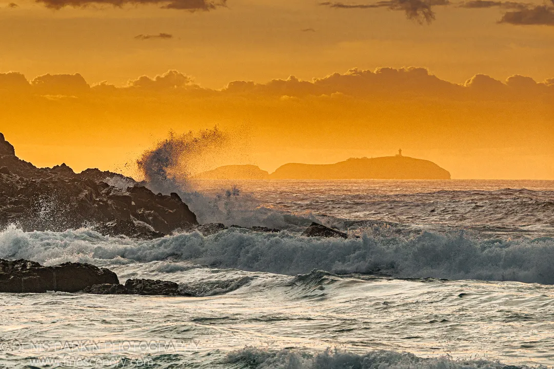 Waves crash against dark basalt rocks before dawn at Coffs Harbor, with a distant lighthouse silhouetted against the orange sky.