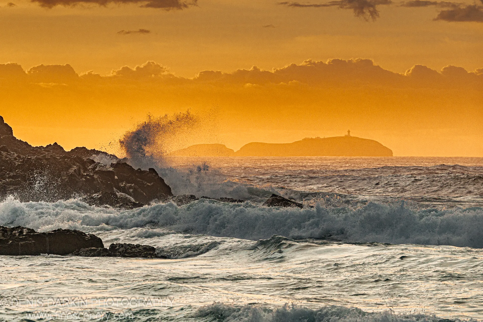 Waves crash against dark basalt rocks before dawn at Coffs Harbor, with a distant lighthouse silhouetted against the orange sky.