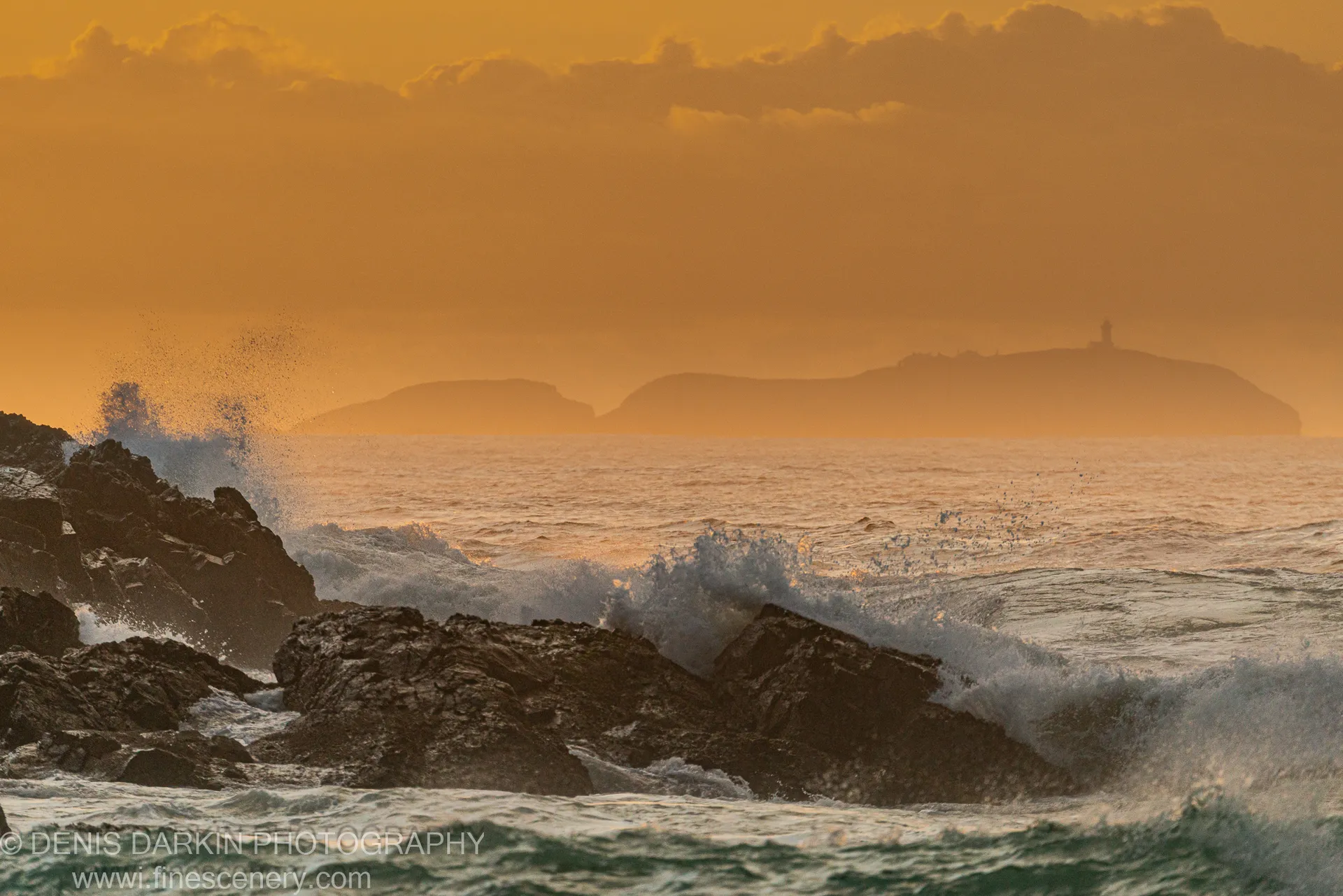 Dark basalt foreground rocks contrast with breaking waves and a warm orange sky, with silhouetted headlands and distant lighthouse on the NSW coast.