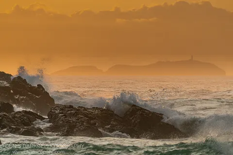 Dark basalt foreground rocks contrast with breaking waves and a warm orange sky, with silhouetted headlands and distant lighthouse on the NSW coast.