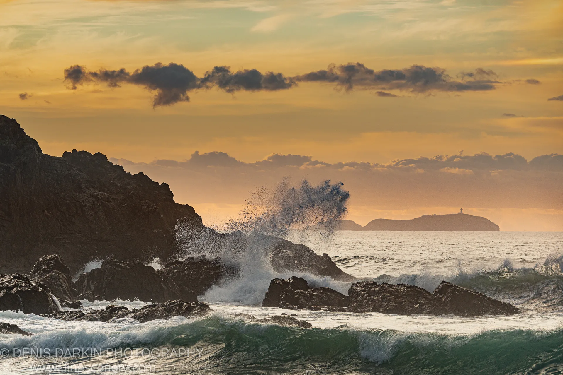 A wave hangs frozen above dark volcanic rocks on the NSW coast at predawn, with a distant headland and lighthouse on the horizon.