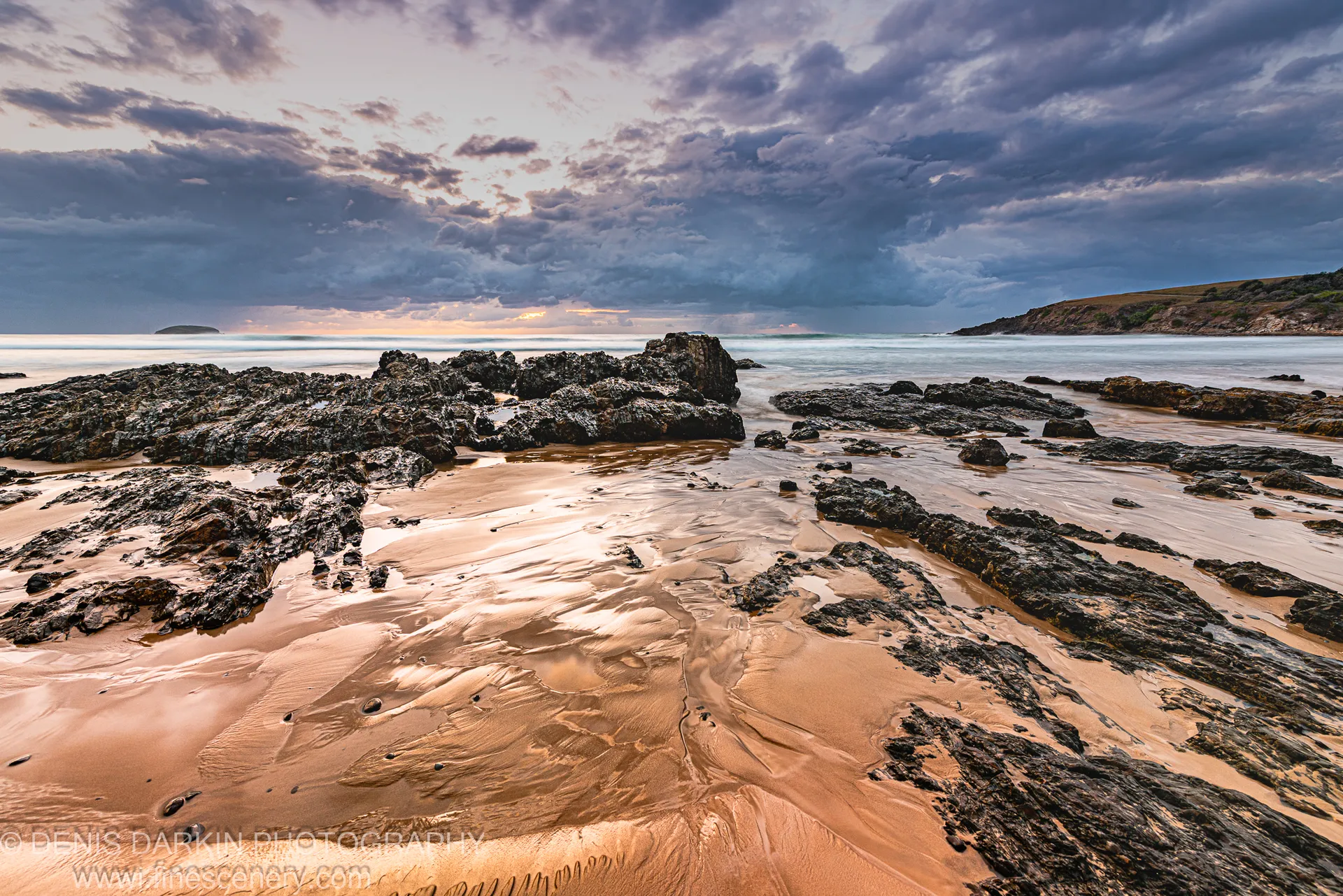 Wet sand ripples on a tidal flat at Coffs Harbor reflect the predawn sky above dark basaltic rock outcrops.