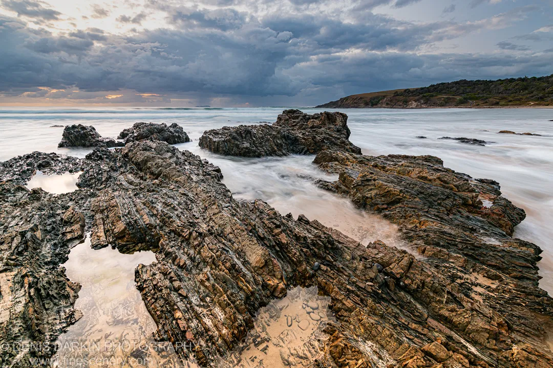 Fractured coastal rock strata at Coffs Harbor with long-exposure surf and a low bank of storm cloud above the headland.