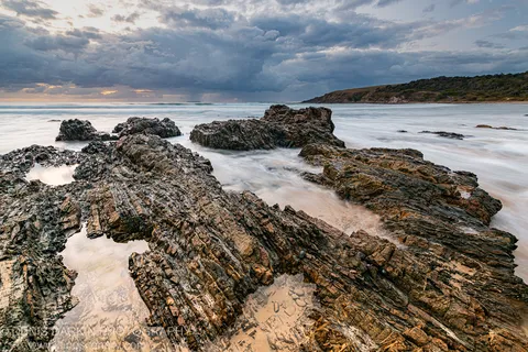 Fractured coastal rock strata at Coffs Harbor with long-exposure surf and a low bank of storm cloud above the headland.