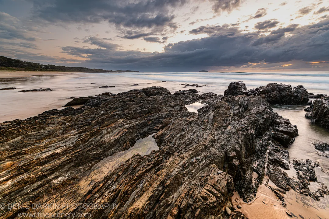 Layered coastal rock formations at Coffs Harbor lead toward a distant island beneath heavy cloud on a spring morning before sunrise.