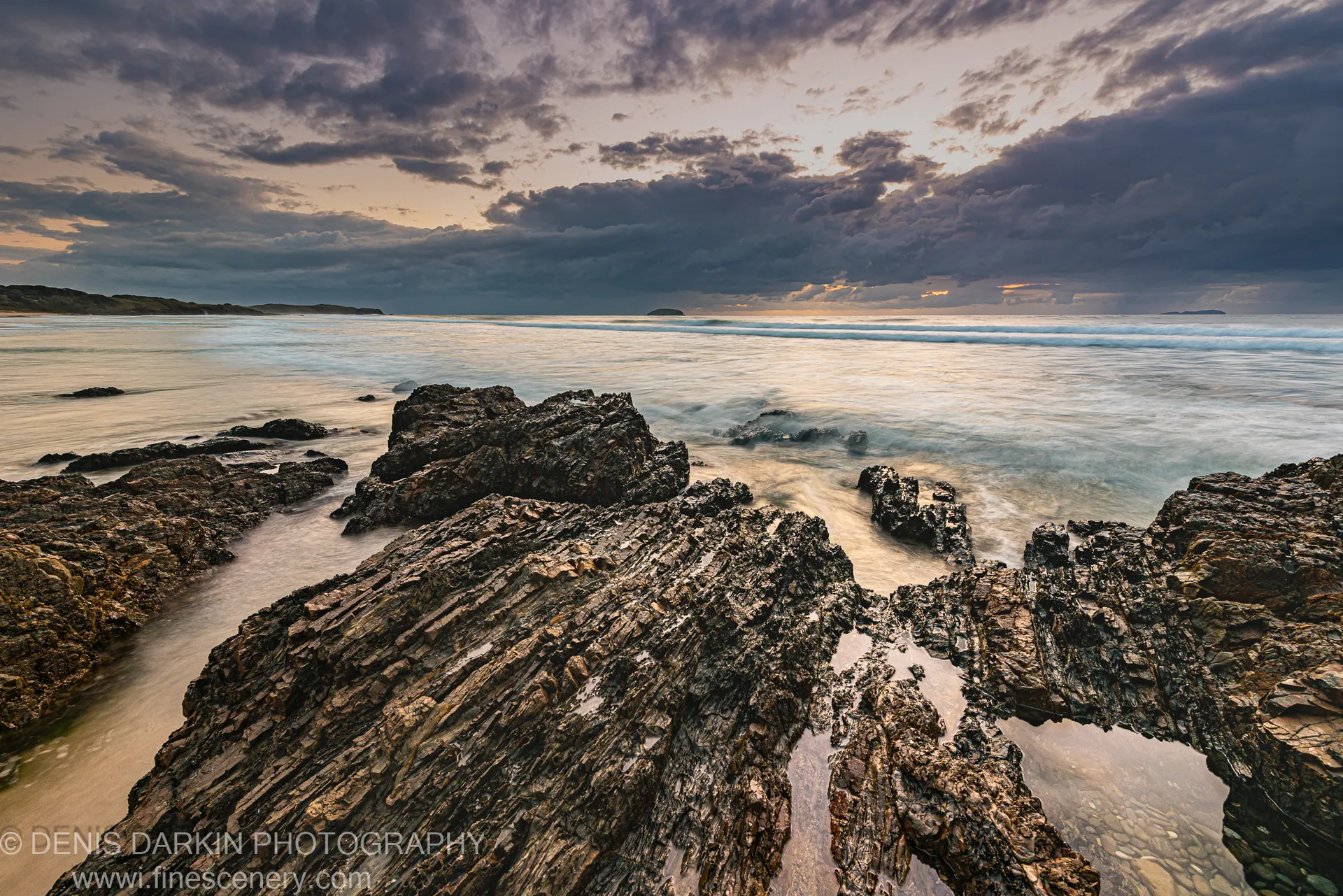 Wet coastal rock shelf at Coffs Harbor with long-exposure waves turned white and a thin warm glow on the horizon below dark cloud.