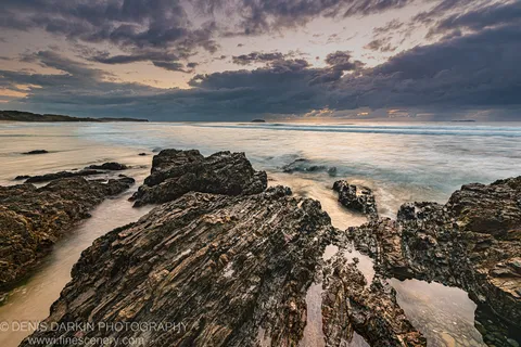 Wet coastal rock shelf at Coffs Harbor with long-exposure waves turned white and a thin warm glow on the horizon below dark cloud.