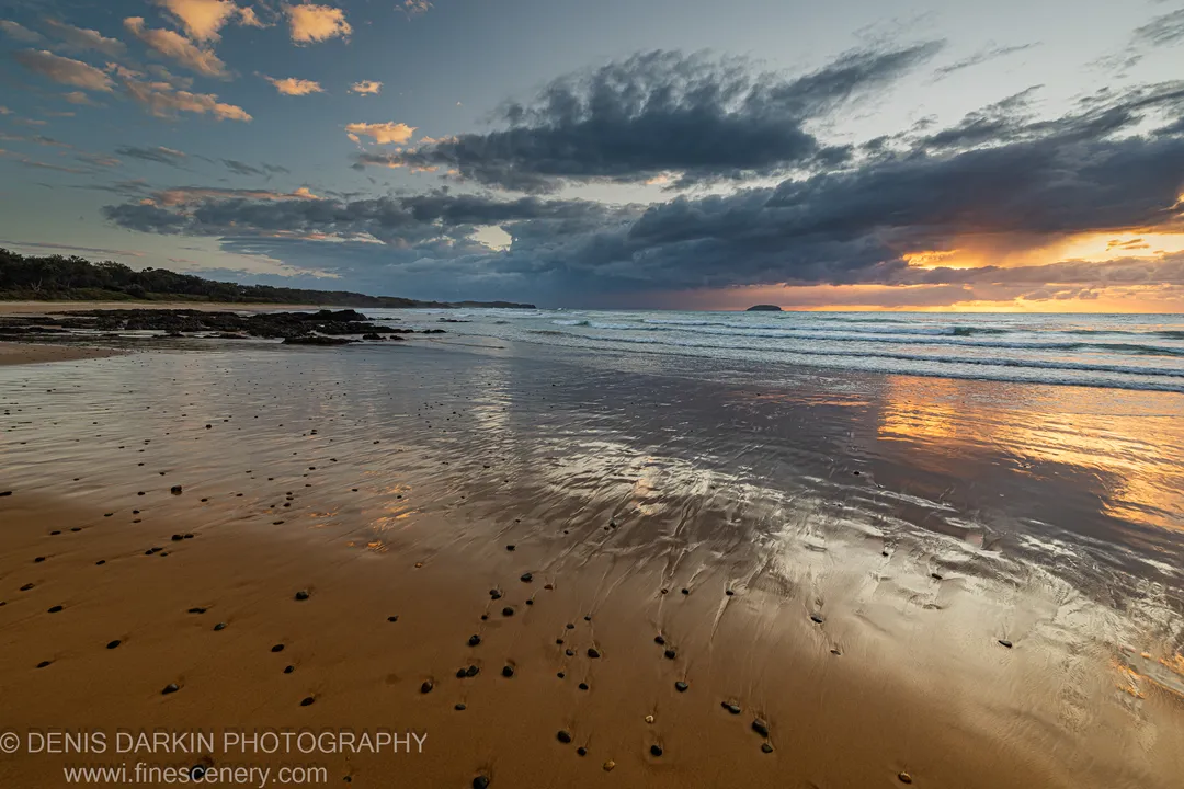 Wet sand and scattered pebbles on a tidal flat at Coffs Harbor below dark storm clouds, with a warm glow near the horizon.