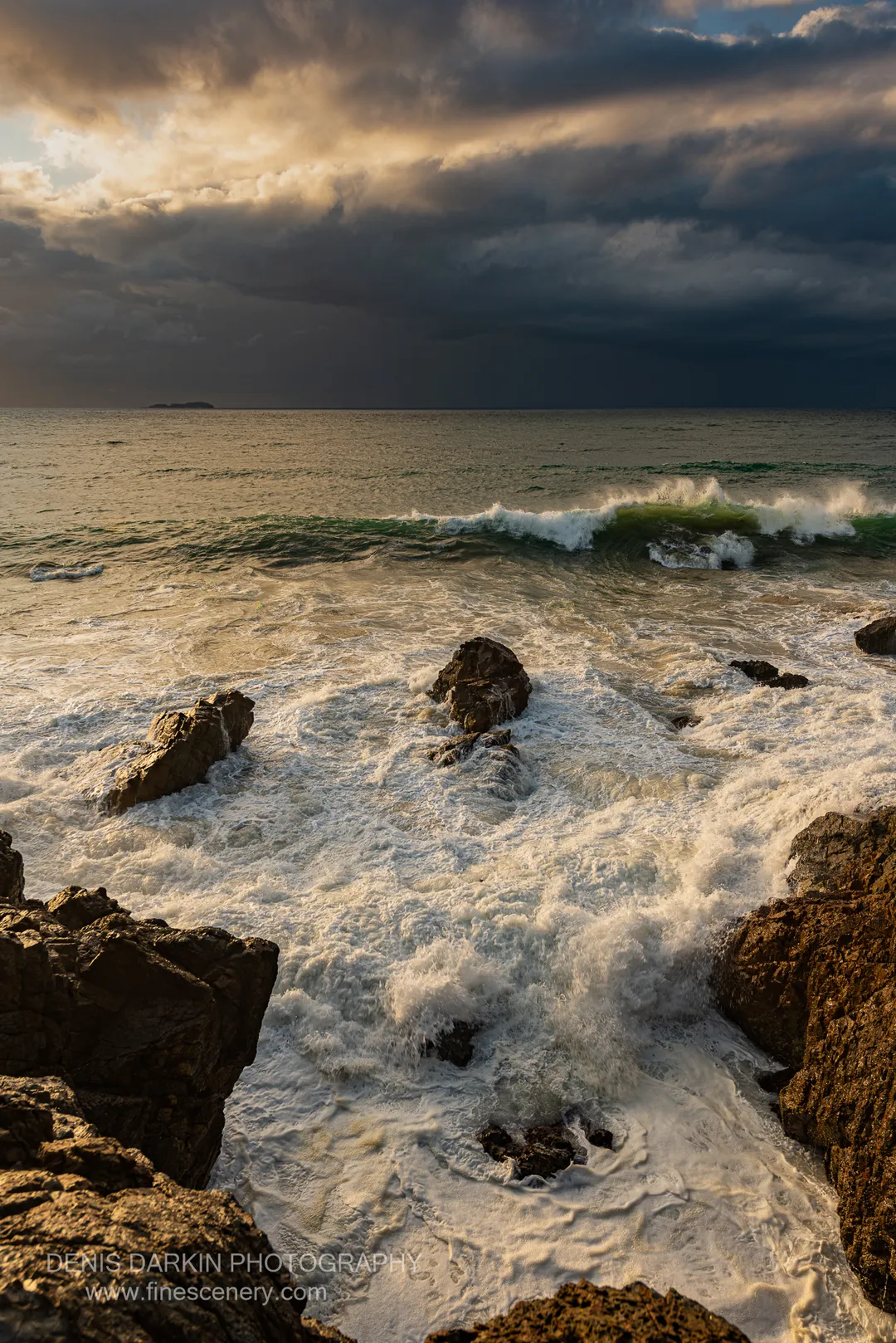 Golden dawn light filters through heavy storm clouds onto wave foam washing over dark coastal rocks at Coffs Harbor.