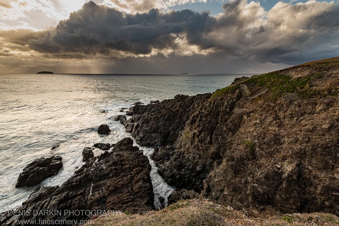 Dark coastal cliffs at Coffs Harbor fall to a calm sea below heavy storm clouds, with warm light along the horizon at dawn.