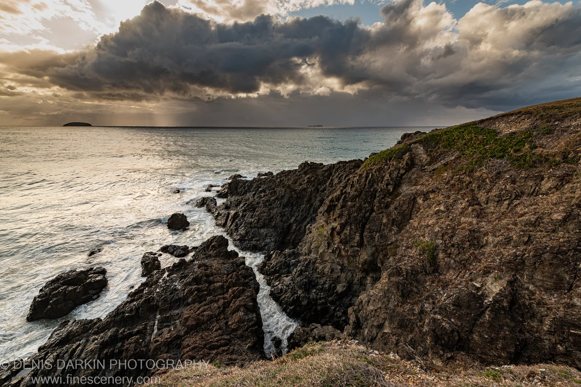 Dark coastal cliffs at Coffs Harbor fall to a calm sea below heavy storm clouds, with warm light along the horizon at dawn.