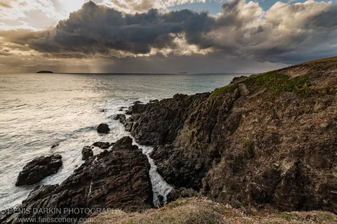 Dark coastal cliffs at Coffs Harbor fall to a calm sea below heavy storm clouds, with warm light along the horizon at dawn.