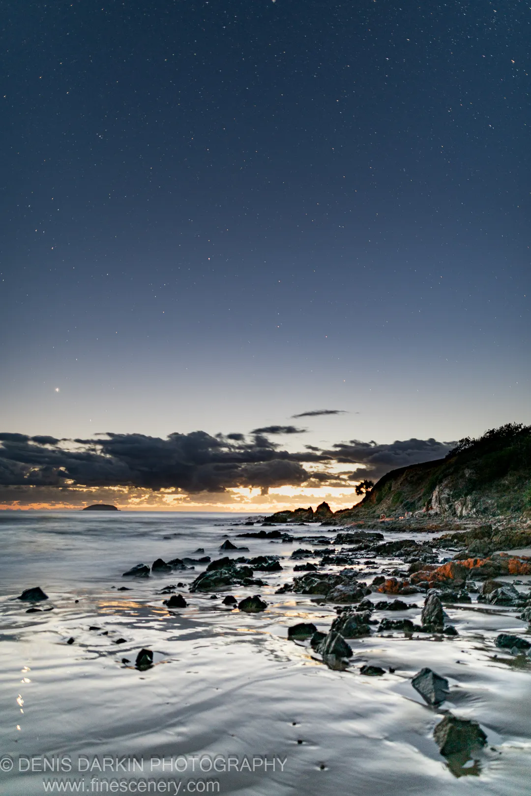 Stars fill the sky above Coffs Harbor as a long exposure captures reflected orange light on wet rocks and still ocean water.