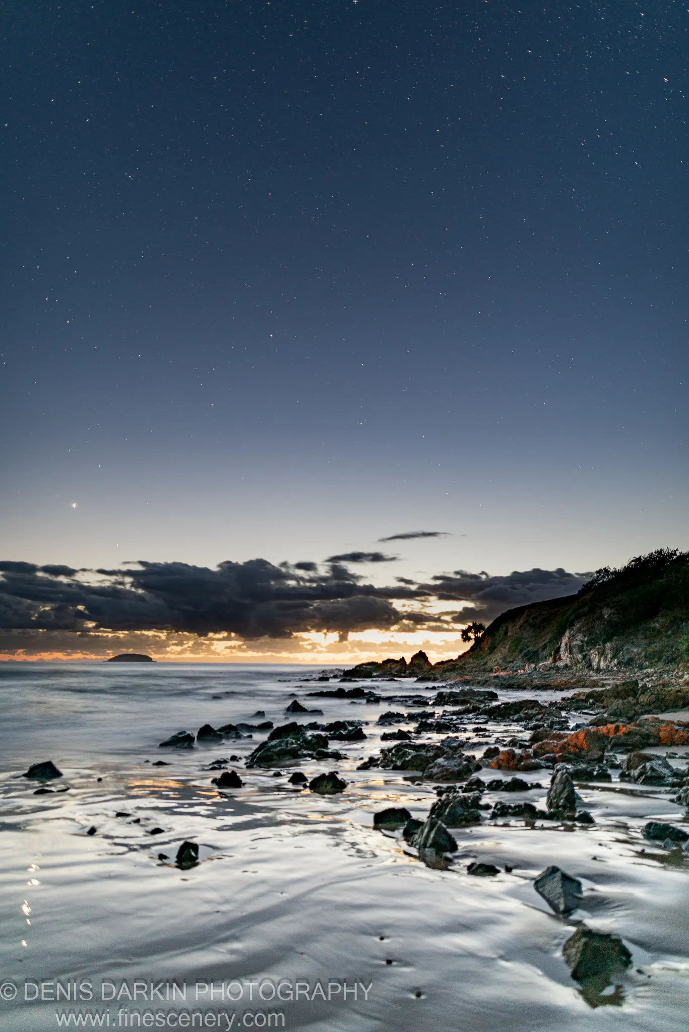 Stars fill the sky above Coffs Harbor as a long exposure captures reflected orange light on wet rocks and still ocean water.