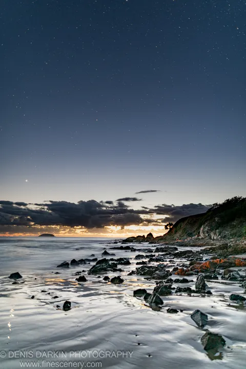 Stars fill the sky above Coffs Harbor as a long exposure captures reflected orange light on wet rocks and still ocean water.