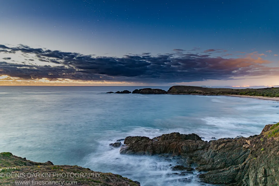 Rugged dark rocks anchor the foreground at Coffs Harbor as the predawn sky shifts from deep blue to warm light at the horizon.