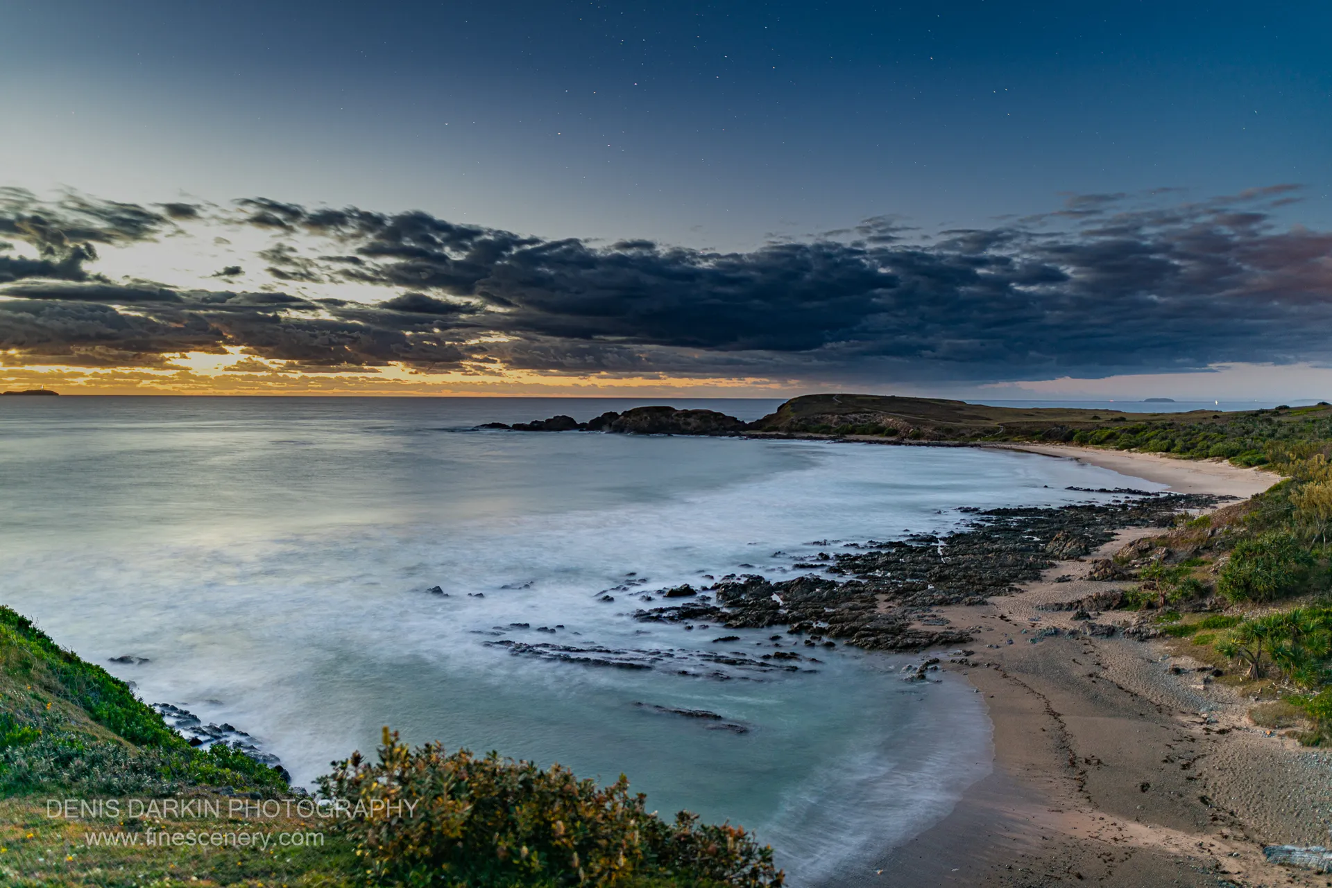 Long-exposure seascape at blue hour near Coffs Harbor, with rocky headland, sandy beach, and golden light along the horizon.