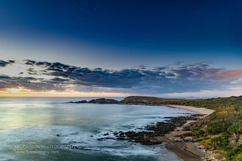 Night sky with stars over a coastal bay at Coffs Harbor, with long-exposure ocean, warm horizon glow, and rocky foreground.