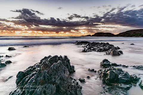 Craggy dark rocks in the foreground of a predawn coastal scene near Coffs Harbor, with smooth long-exposure water and golden horizon light.