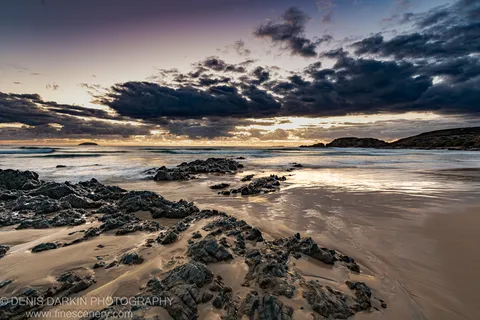 Rugged tidal rocks and wet sand reflecting a golden horizon glow at Coffs Harbor, with storm clouds overhead and long-exposure ocean waves.