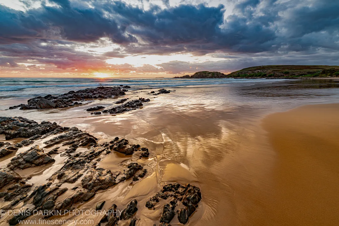 Wet sand reflecting golden sunrise light at Coffs Harbor, with basalt rock formations and heavy storm clouds over the headland.