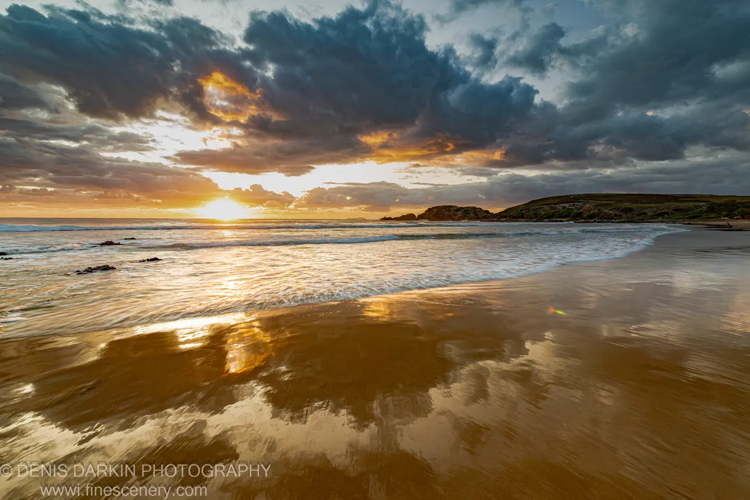 Wide coastal beach at predawn near Coffs Harbor, where sunlight breaks through storm clouds and casts a golden path across the wet sand.
