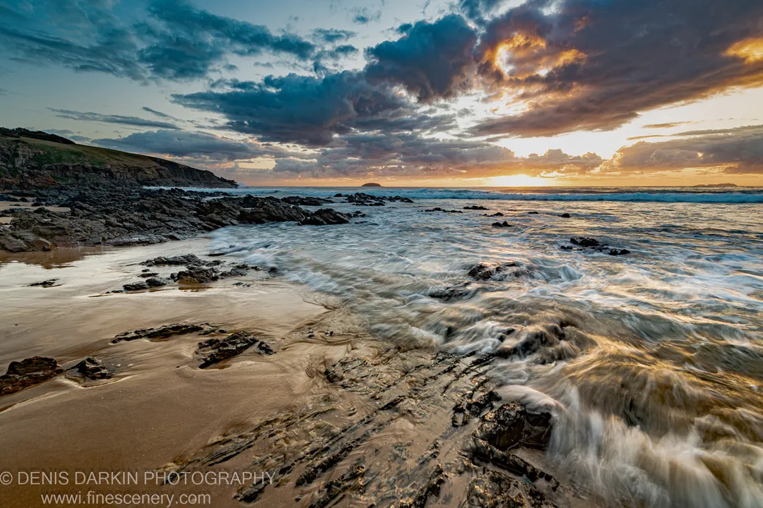 Rocky beach at predawn near Coffs Harbor, with motion-blurred surf and a sky ranging from deep blue to orange toward the headland.