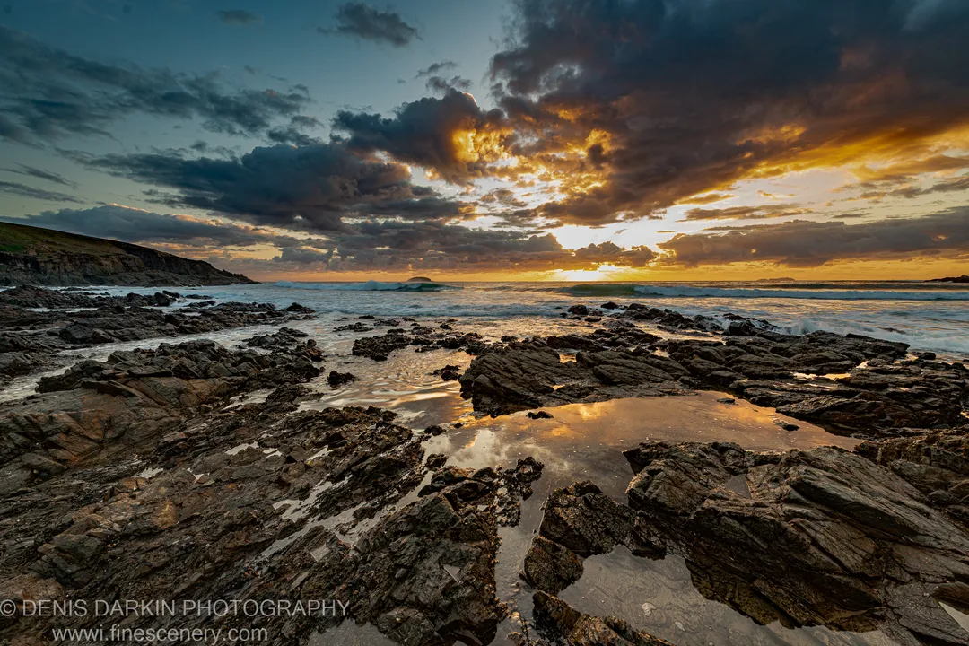 Rough dark rock platform at Coffs Harbor with tidal pools reflecting golden sunrise light breaking through heavy storm clouds.
