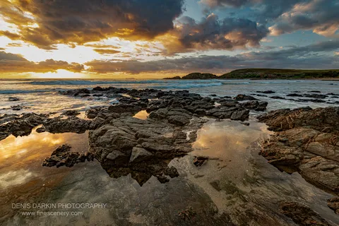 Shallow tidal pools on a rocky shoreline at Coffs Harbor reflect the amber sky as sunrise light breaks through heavy storm clouds.