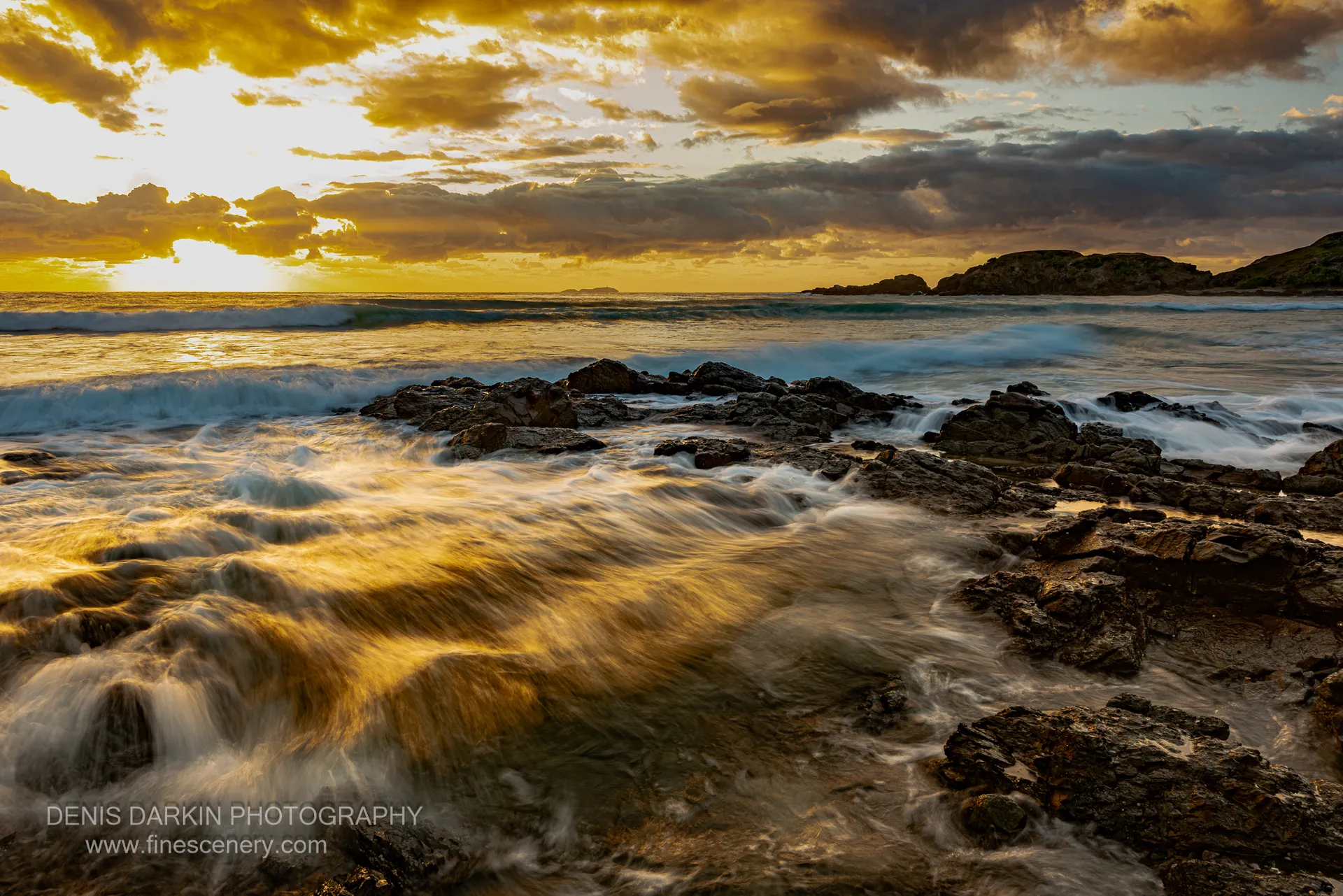 Motion-blurred waves wash over dark rocks on the New South Wales coast before sunrise, under a sky shifting from amber to grey.