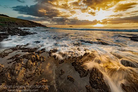 Long exposure waves flow across a wet basalt coastal platform at Coffs Harbor, lit by warm predawn glow beneath textured cumulus clouds.
