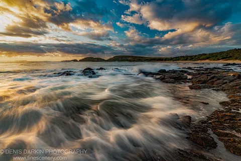 Motion-blurred waves pass over tidal rocks at predawn, with grassy headlands visible across the bay under blue and gold cloud.