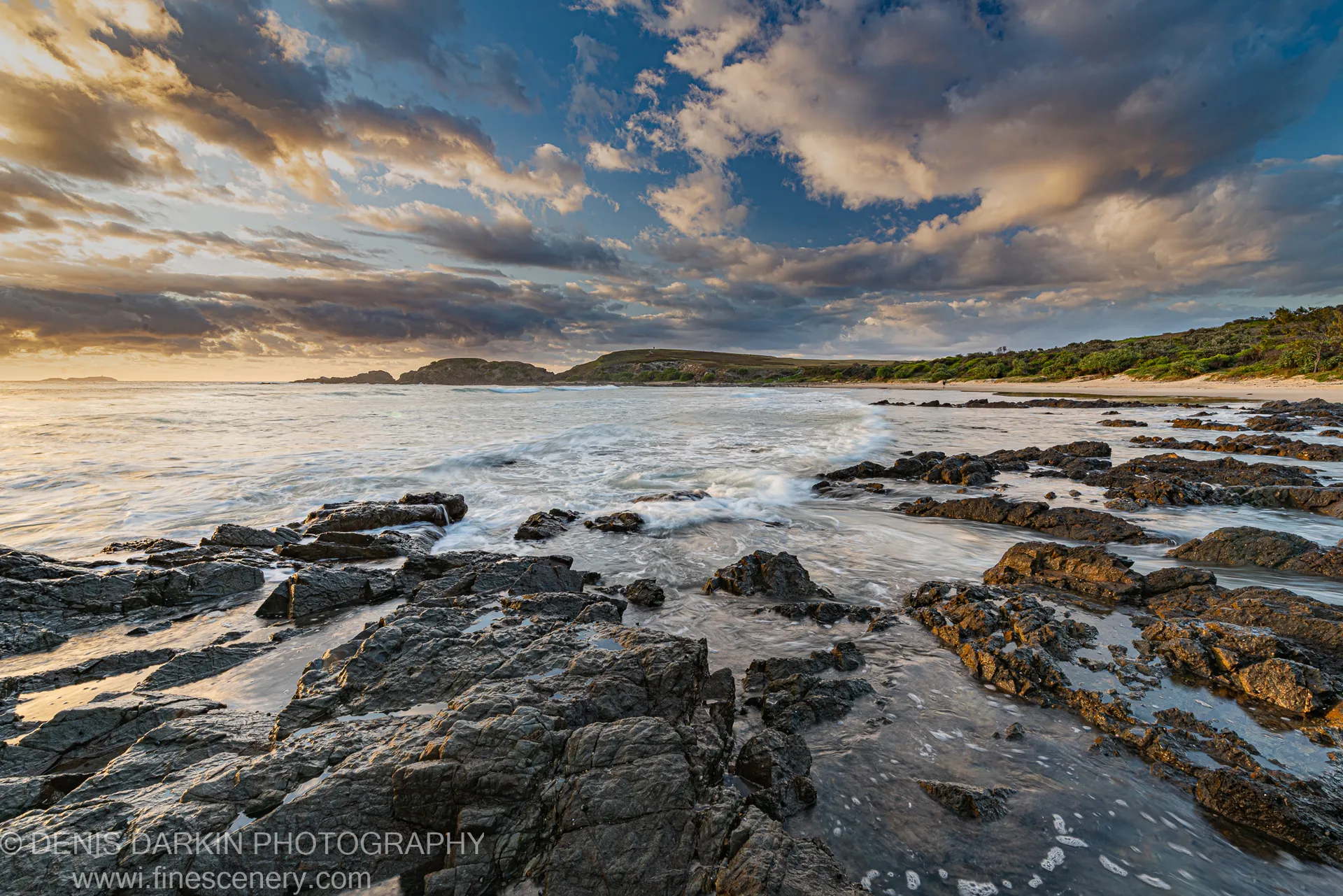 Foam-washed basalt rocks occupy the foreground of a curving bay at Coffs Harbor, with silhouetted headlands against an early spring sky.