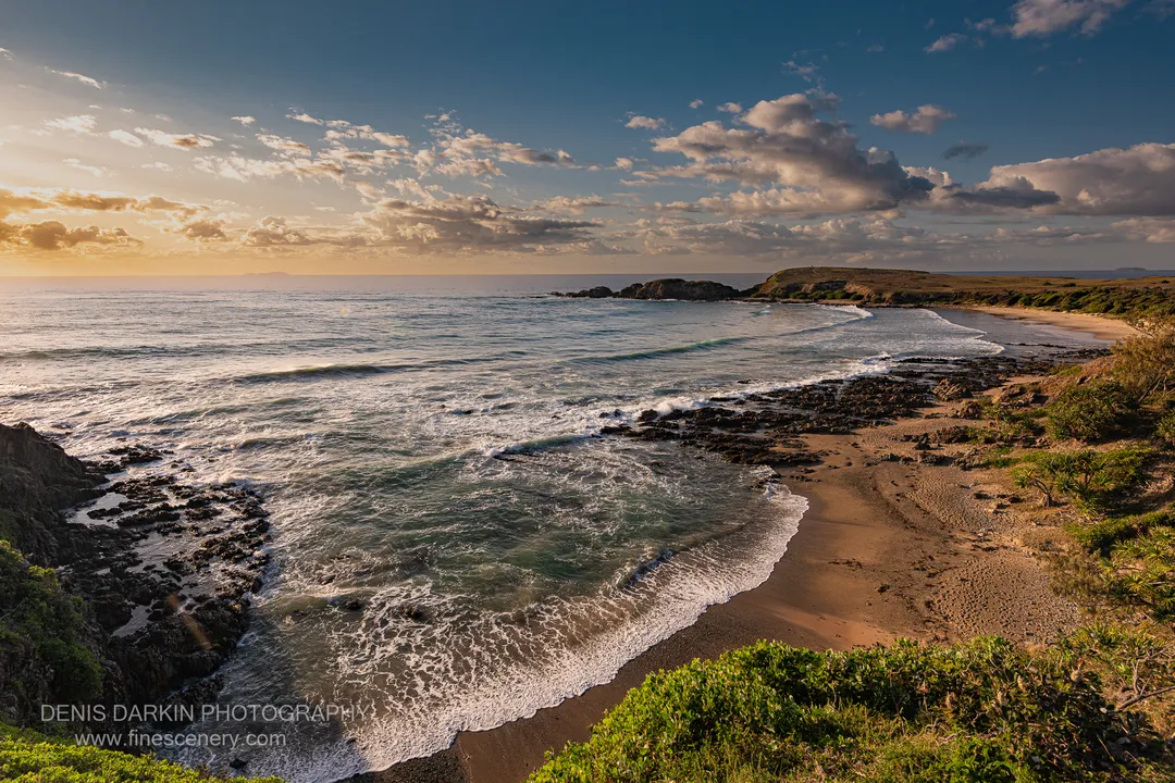 Wave foam creates patterns against dark rocks and smooth sand on the NSW coast at dawn, with coastal scrub at the water's edge.