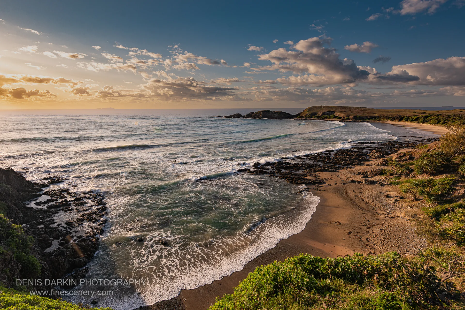 Wave foam creates patterns against dark rocks and smooth sand on the NSW coast at dawn, with coastal scrub at the water's edge.