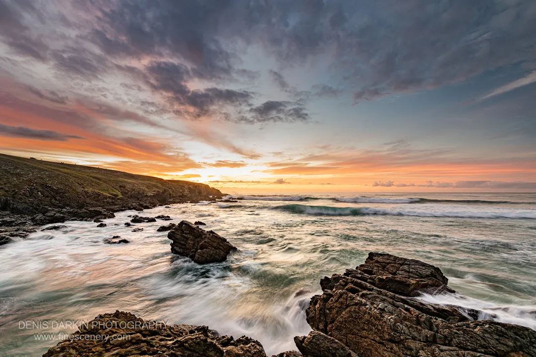 A deep blue sky fades to orange above a grassy headland on the NSW coast at predawn, with angular rocks in the foreground.