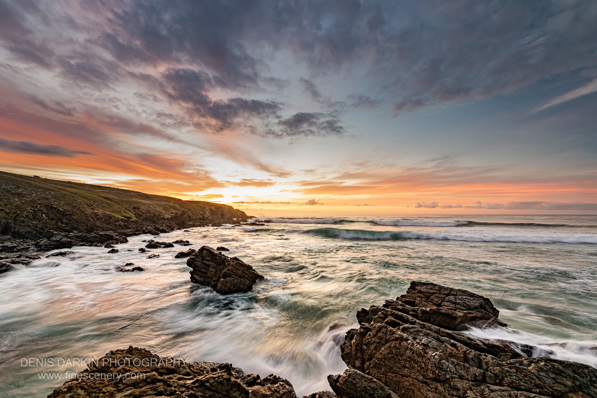 A deep blue sky fades to orange above a grassy headland on the NSW coast at predawn, with angular rocks in the foreground.
