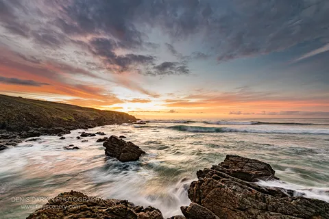 A deep blue sky fades to orange above a grassy headland on the NSW coast at predawn, with angular rocks in the foreground.