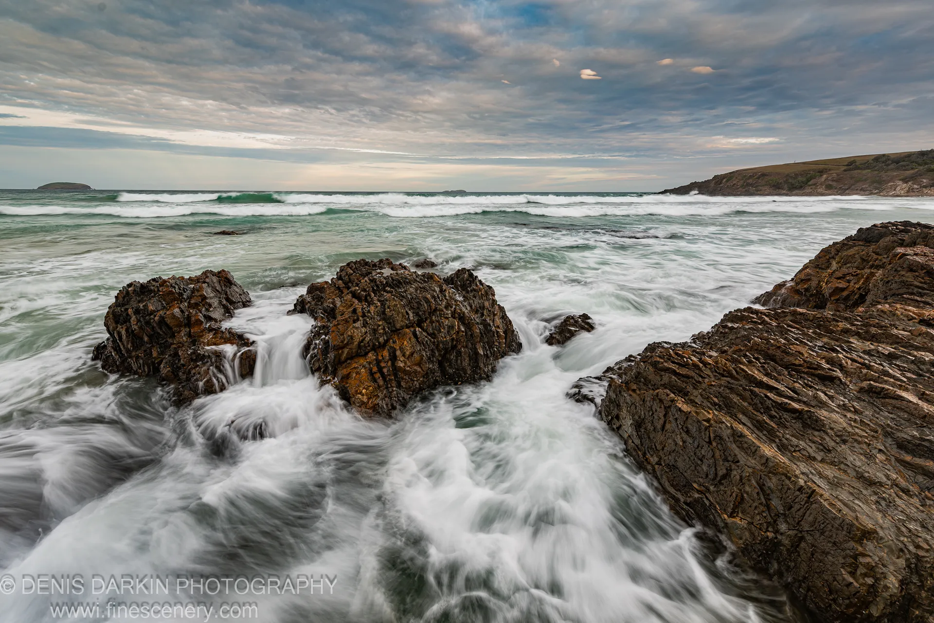 Long-exposure waves break over a stratified rock shelf on the New South Wales coast, blurred surf against layered stone under a soft grey sky.