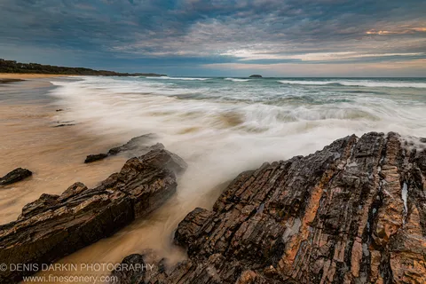 Dark layered sandstone in the foreground with long-exposure surf washing a sandy beach below a streaked evening sky on the New South Wales coast.