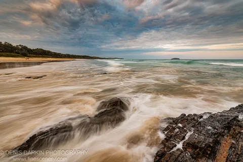 Long-exposure surf over dark layered rocks in the foreground of a curved Coffs Harbor beach, receding tide pooled on the sand.