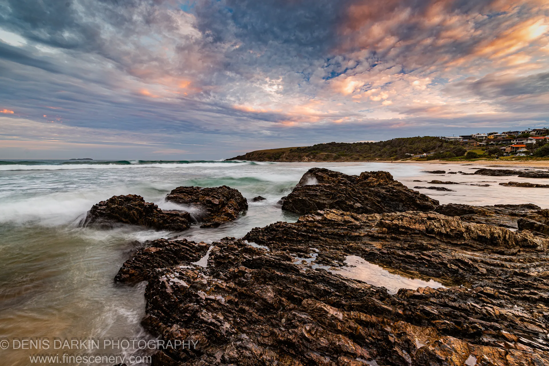 Dark volcanic rock platform in the foreground with long-exposure surf below a pink and orange dusk sky on the New South Wales coast.