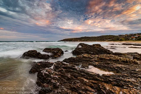 Dark volcanic rock platform in the foreground with long-exposure surf below a pink and orange dusk sky on the New South Wales coast.